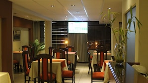 Un comedor de hotel con mesas y sillas, una pantalla grande mostrando contenido y una planta en maceta.