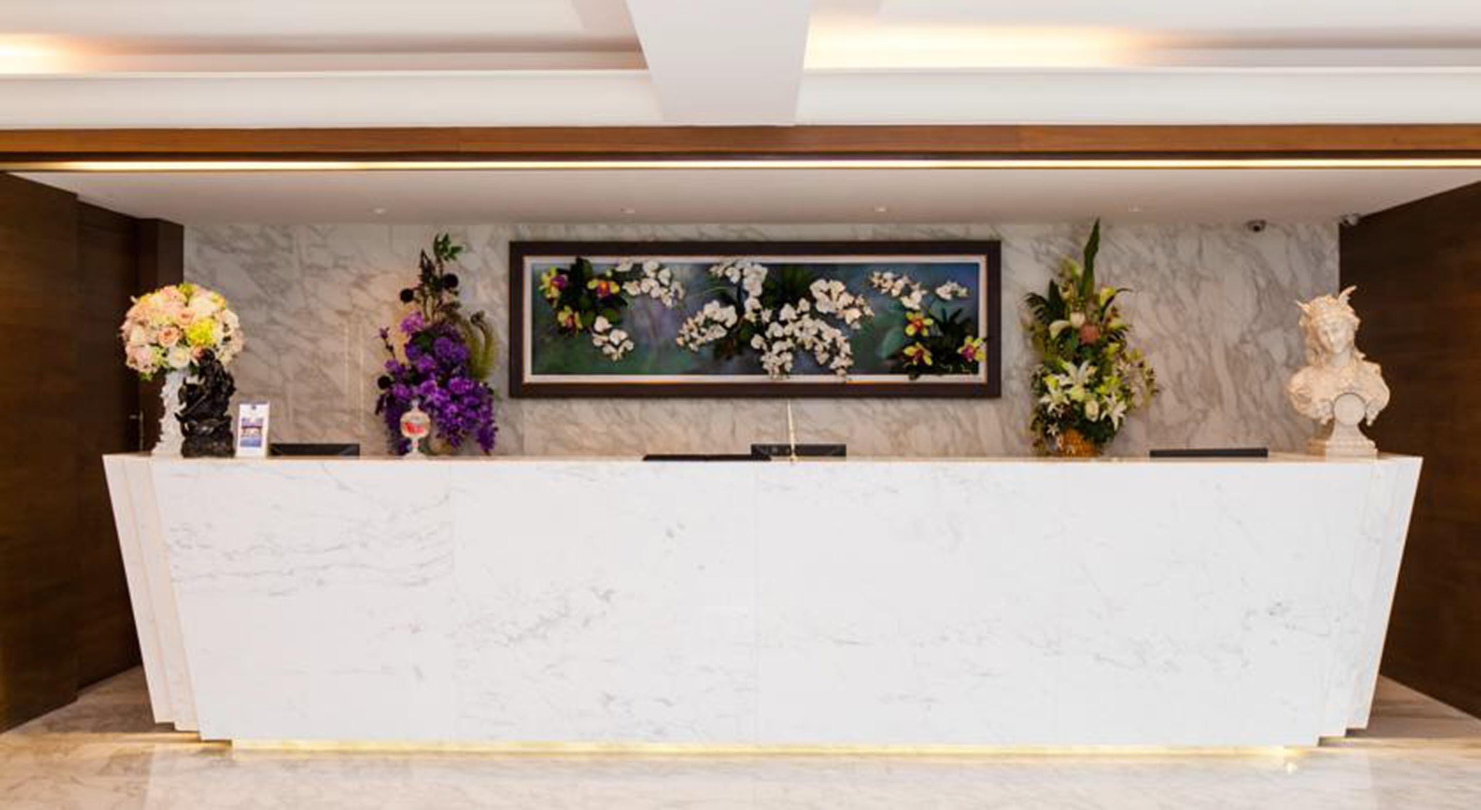 Un comptoir d’accueil surmonté d’une plaque de marbre, agrémenté de part et d’autre de compositions florales et d’un tableau floral encadré.