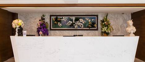 Un comptoir d’accueil surmonté d’une plaque de marbre, agrémenté de part et d’autre de compositions florales et d’un tableau floral encadré.
