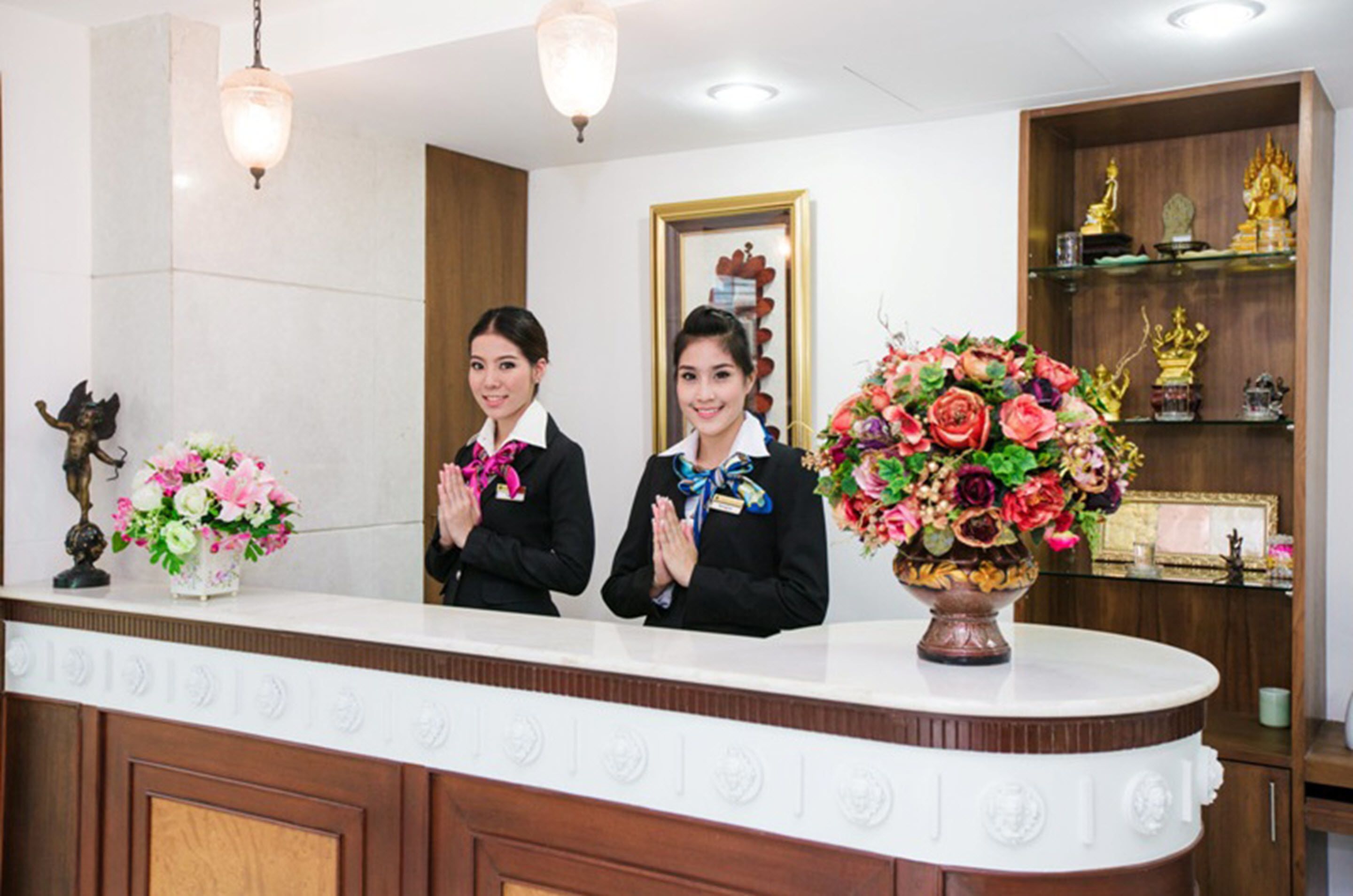 Una recepción con dos empleados, un arreglo floral y un mueble de madera con estatuas.