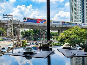 A dining area with a view of a train passing over an elevated structure, a table with a cup and saucer, and a vase with flowers.
