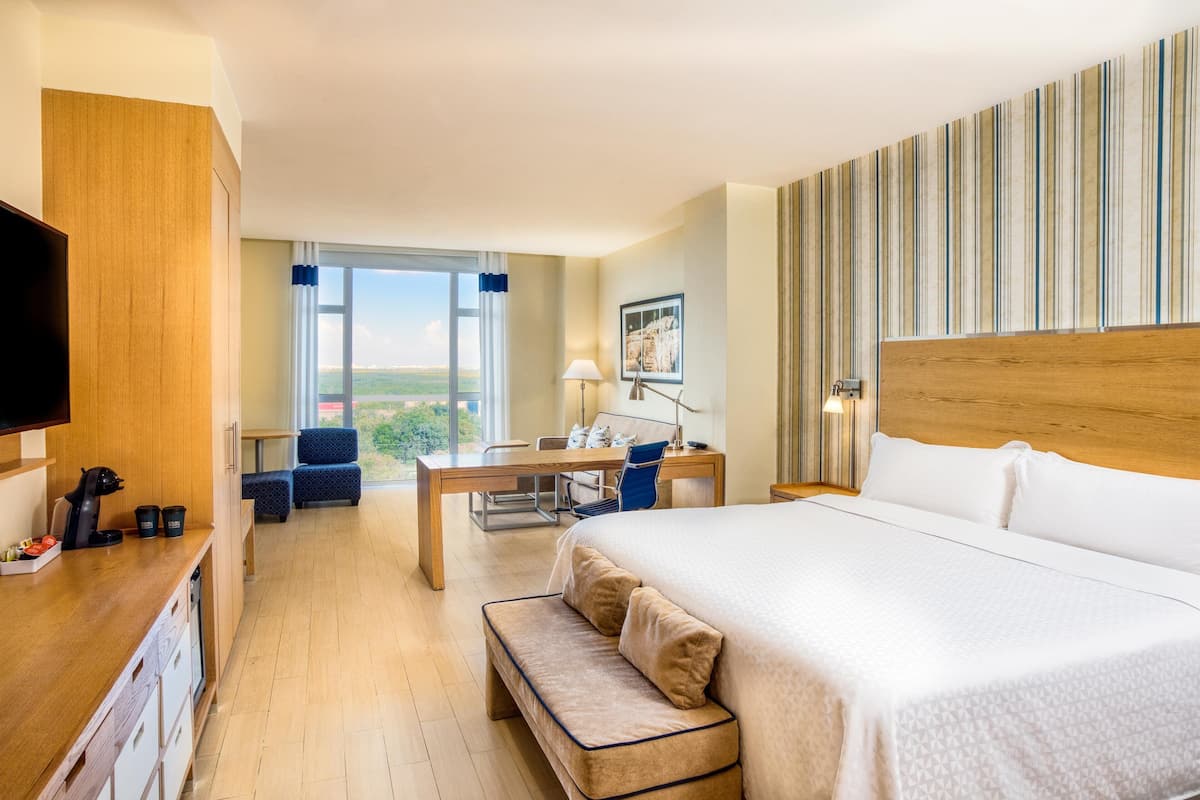 A hotel room with a large bed, a desk with a chair, a bench, a TV, and a window with a view.