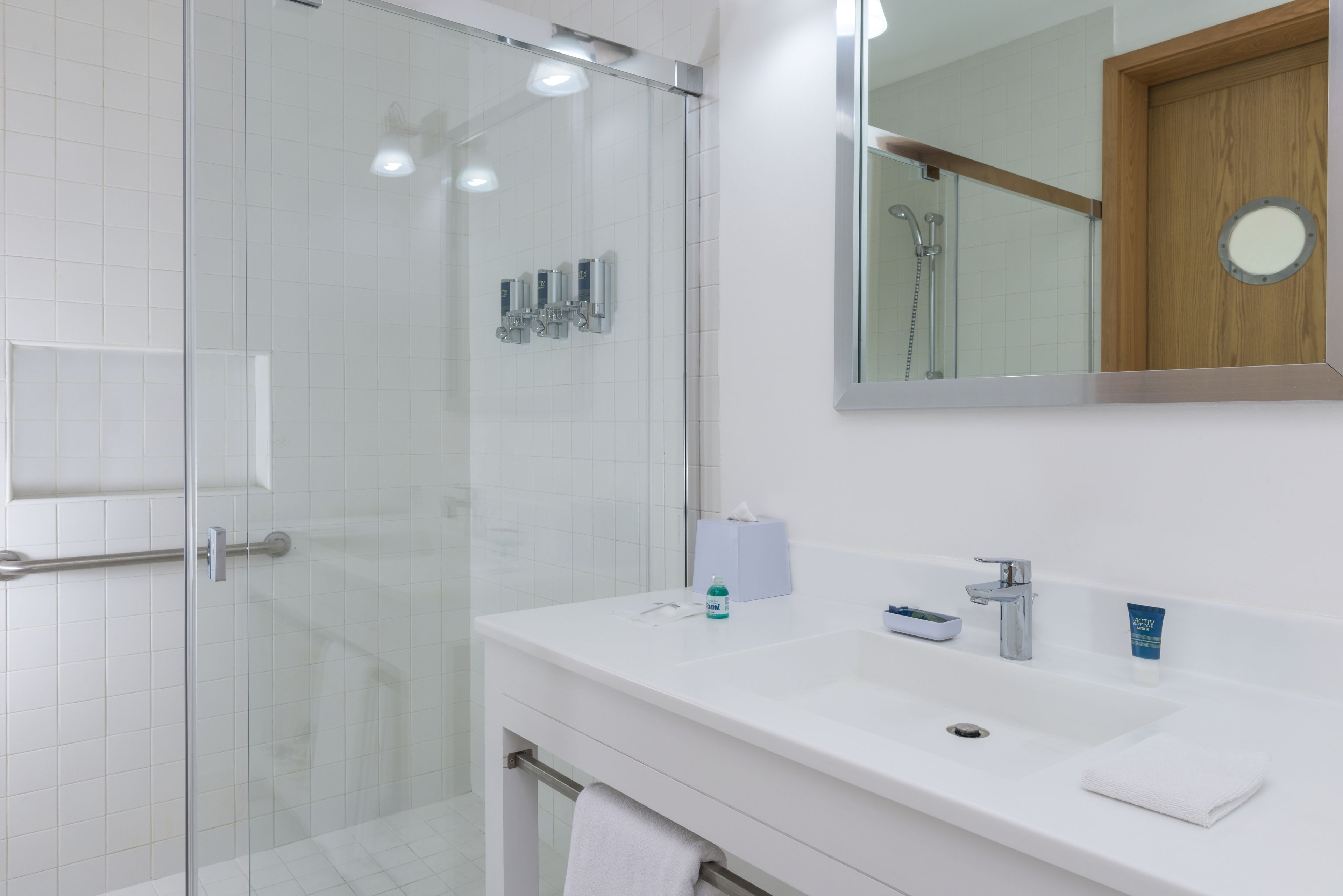 Une salle de bain moderne avec une douche à l’italienne en verre, un lavabo blanc avec un miroir au-dessus, et une porte en bois.