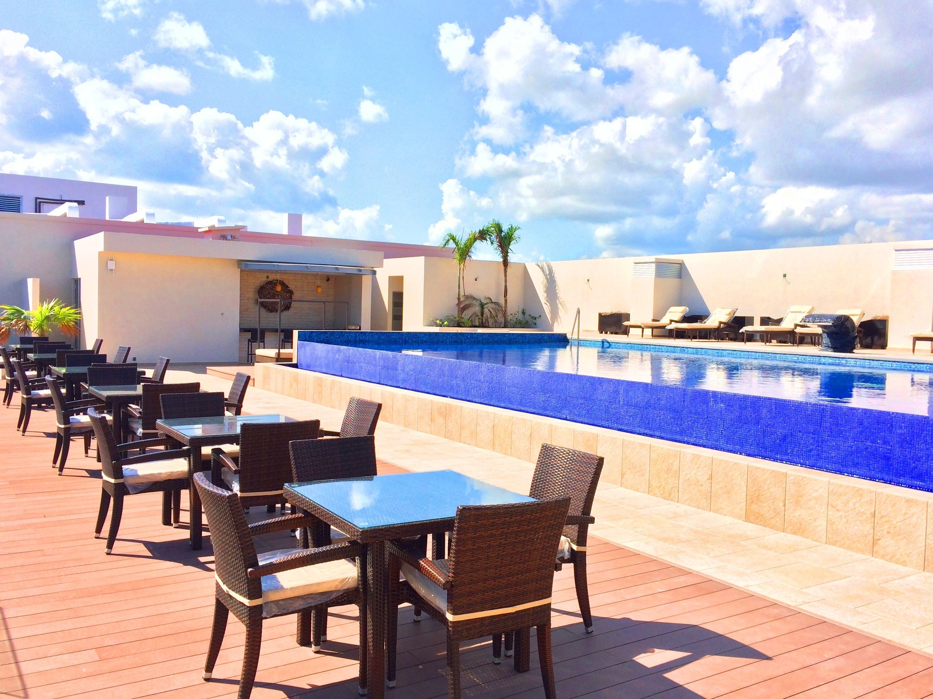 Un espace extérieur avec une piscine, des chaises longues et des tables, un bar, et un ciel d’un bleu éclatant.