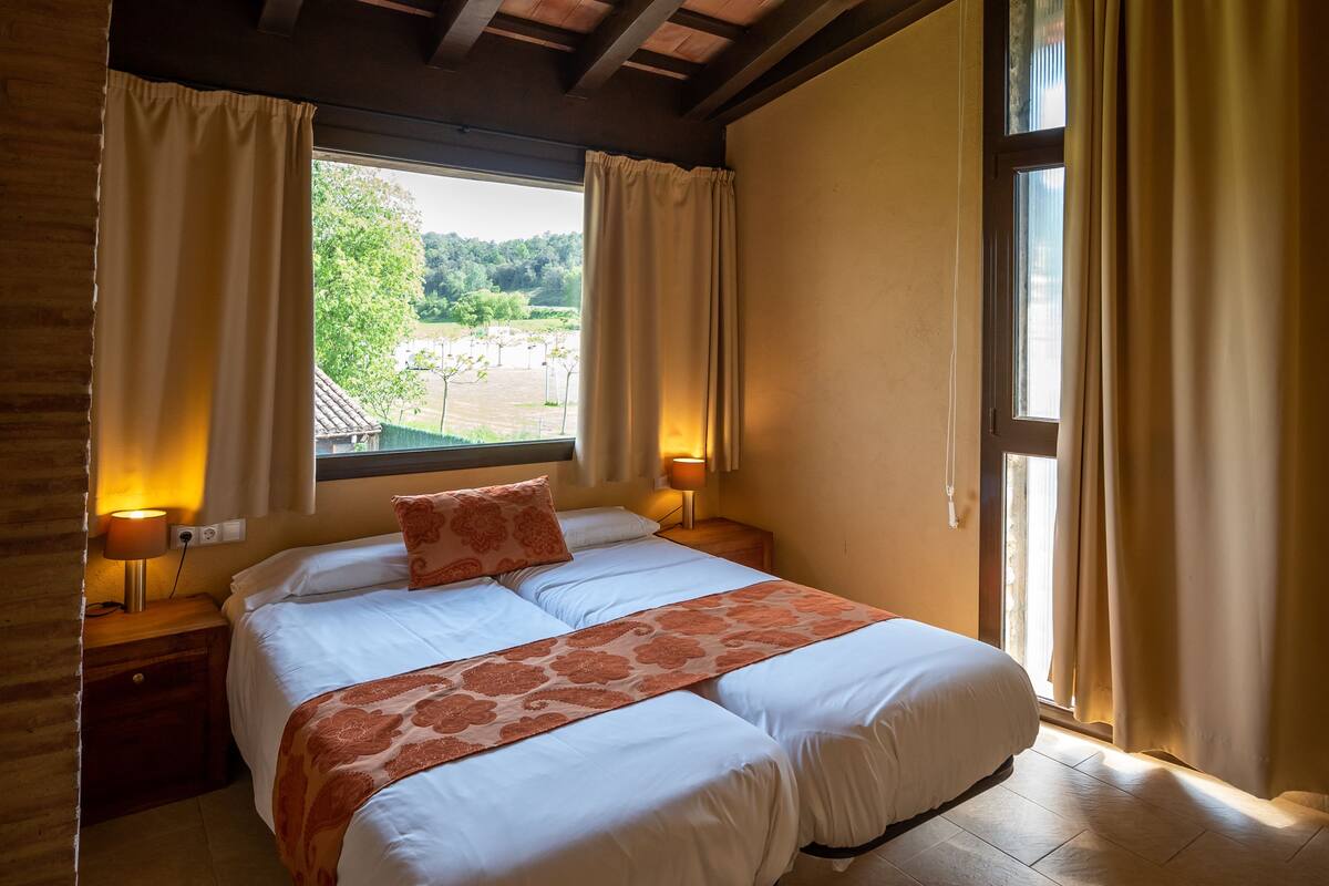 A bedroom with a large bed, wooden ceiling beams, a window with curtains, and a hanging light fixture.