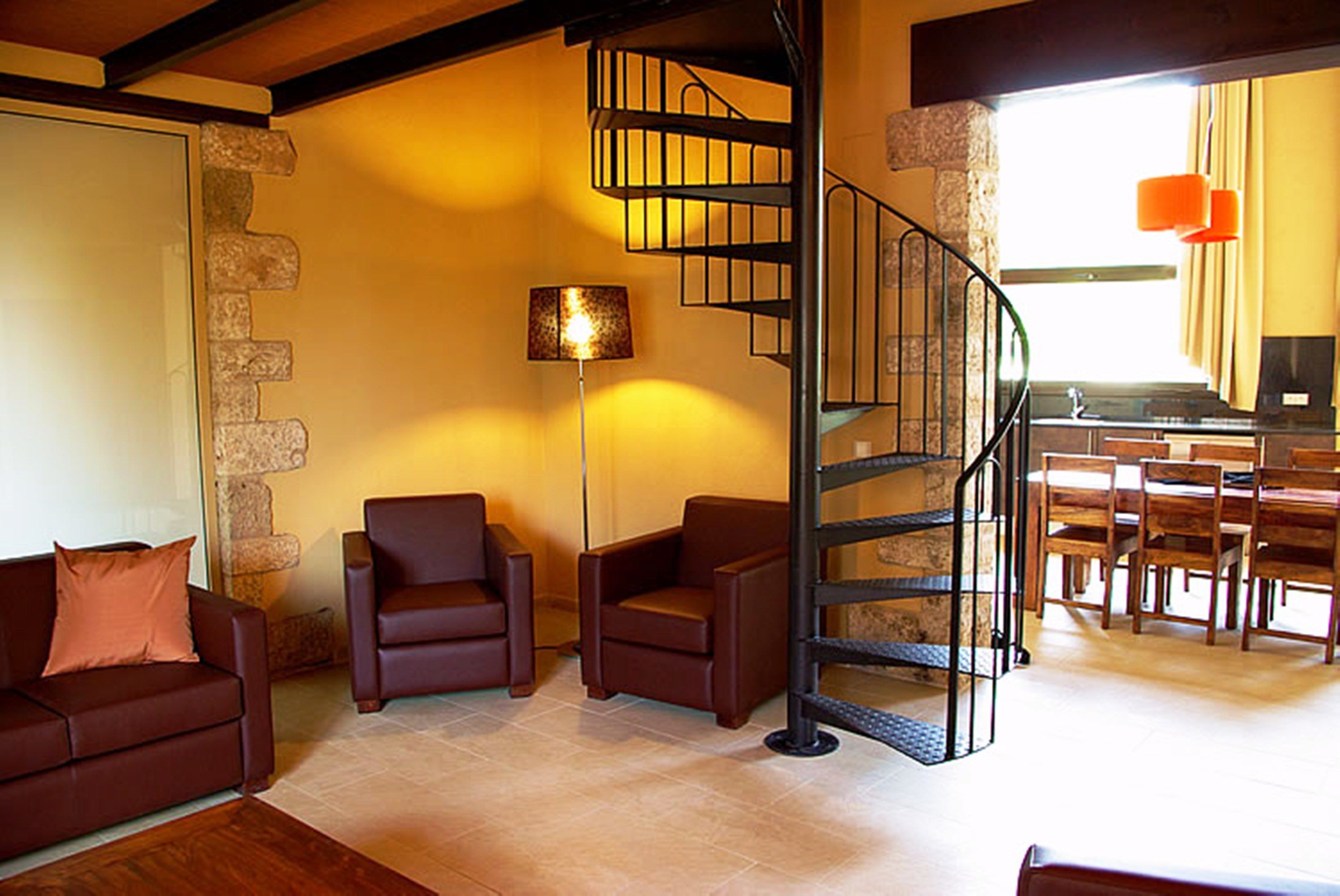 Una sala de estar con un sofá de cuero marrón, dos sillones, una lámpara de pie y una escalera en espiral que conduce a una zona de cocina.