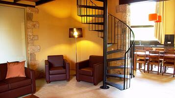 Una sala de estar con un sofá de cuero marrón, dos sillones, una lámpara de pie y una escalera en espiral que conduce a una zona de cocina.