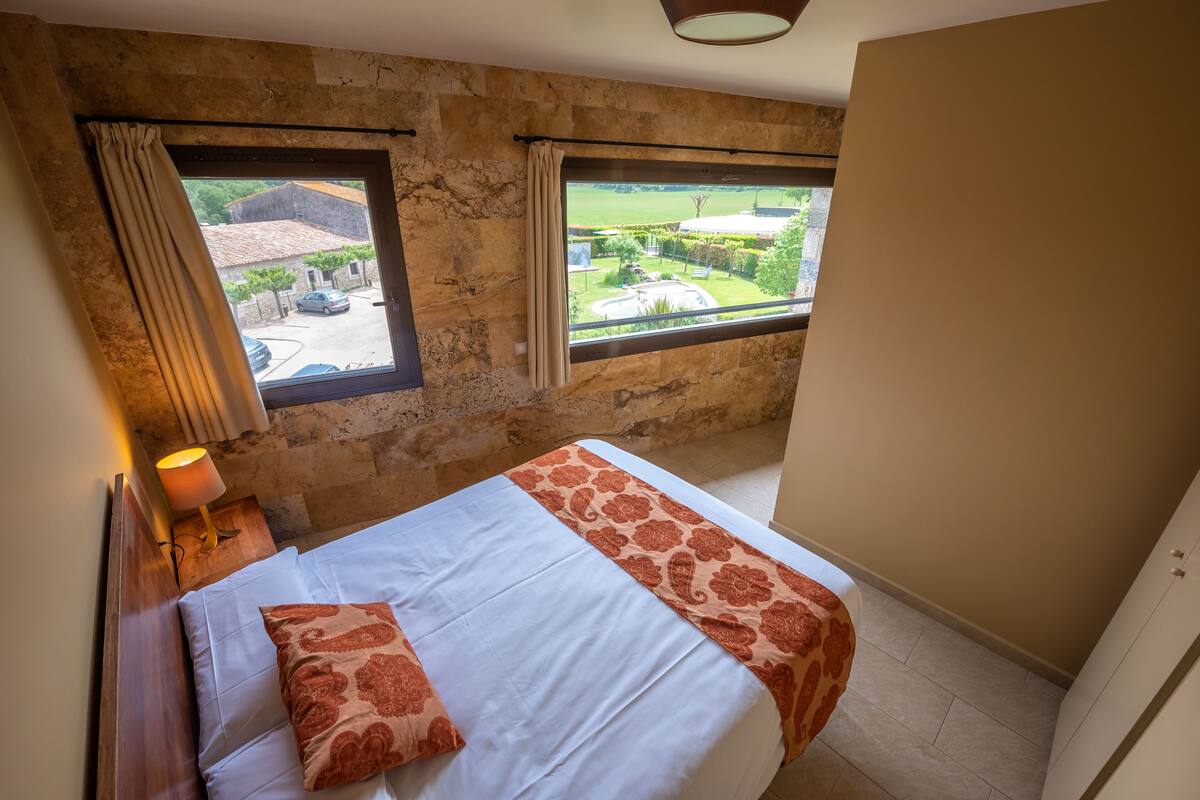 A bedroom with a bed, a nightstand with a lamp, a window with curtains, and a view of a courtyard.