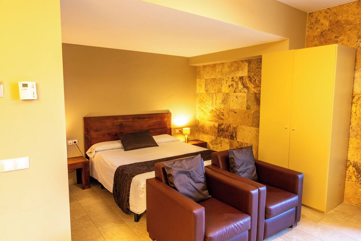 A hotel room with a bed, a wooden headboard, a nightstand, a lamp, a wardrobe, and two leather armchairs.
