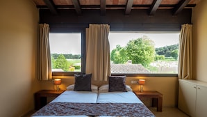 A bedroom with a bed, two bedside tables with lamps, a window with curtains, and a view of greenery and a building outside. - L'Arcada de Fares (Sant Ferriol)