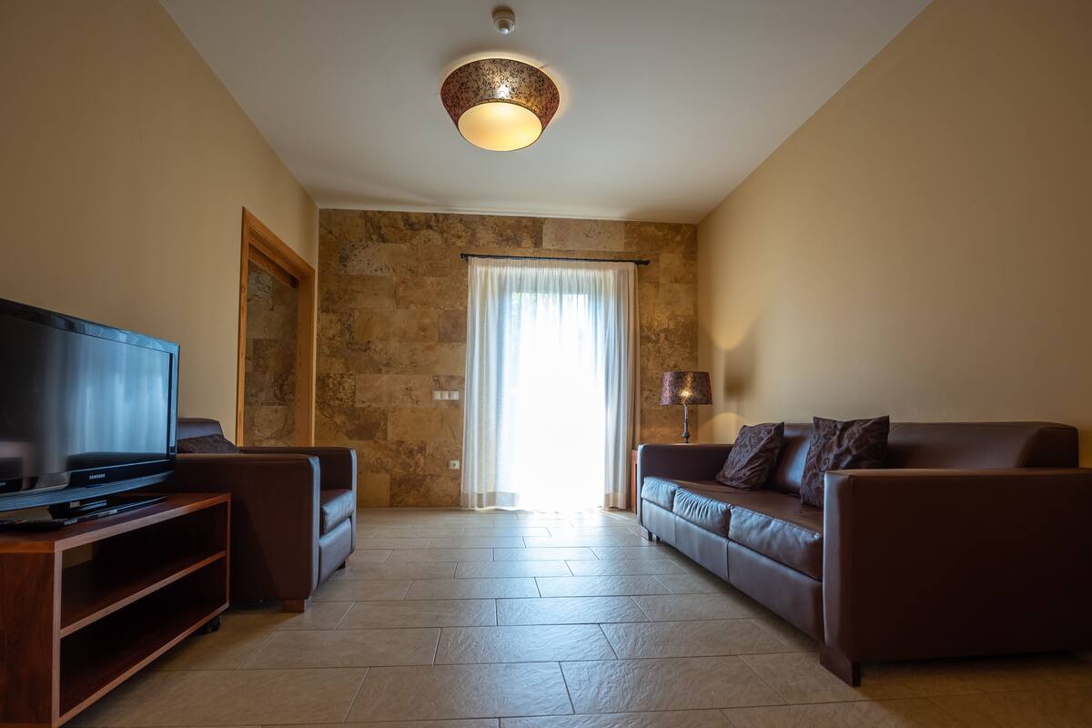 A living room with a flat-screen TV on a wooden stand, a leather sofa, a lamp, and a stone wall.