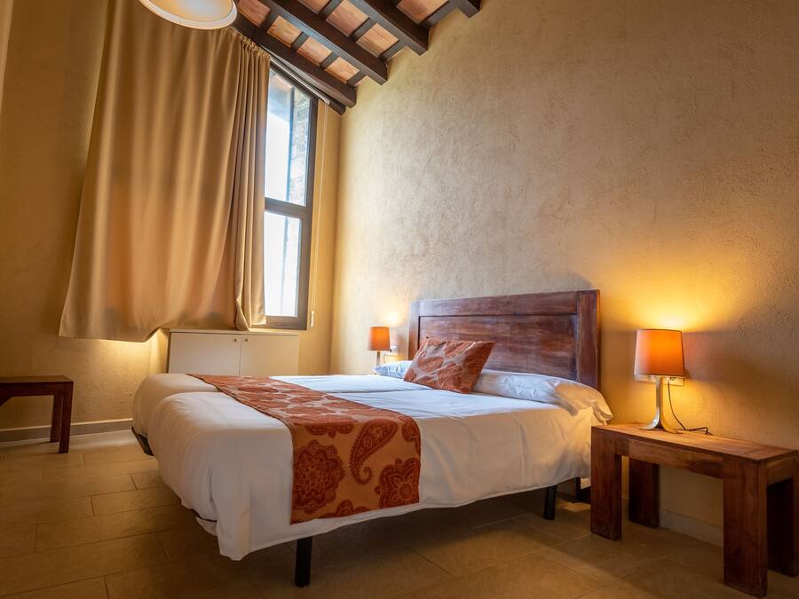 Ein Schlafzimmer mit Holzboden, einem Bett mit gemustertem Überwurf, einem Nachttischchen mit Lampe und einem Fenster mit Vorhängen.