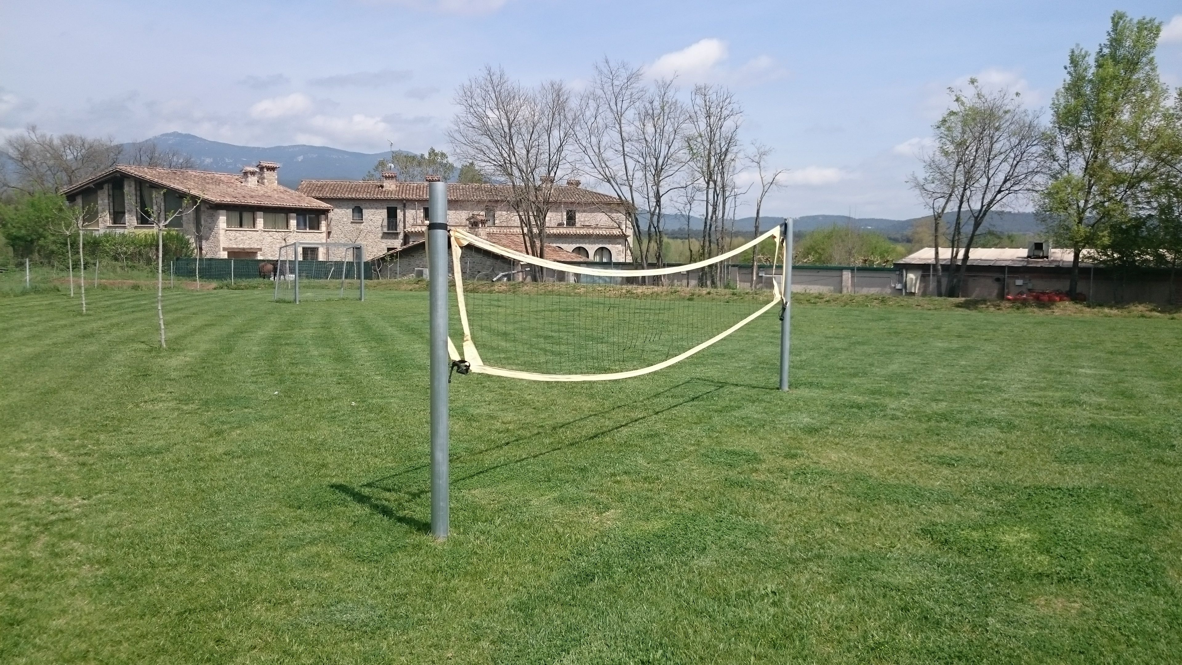A grassy field with a volleyball net in the foreground, a stone building with a tiled roof in the background, and a mountainous landscape.