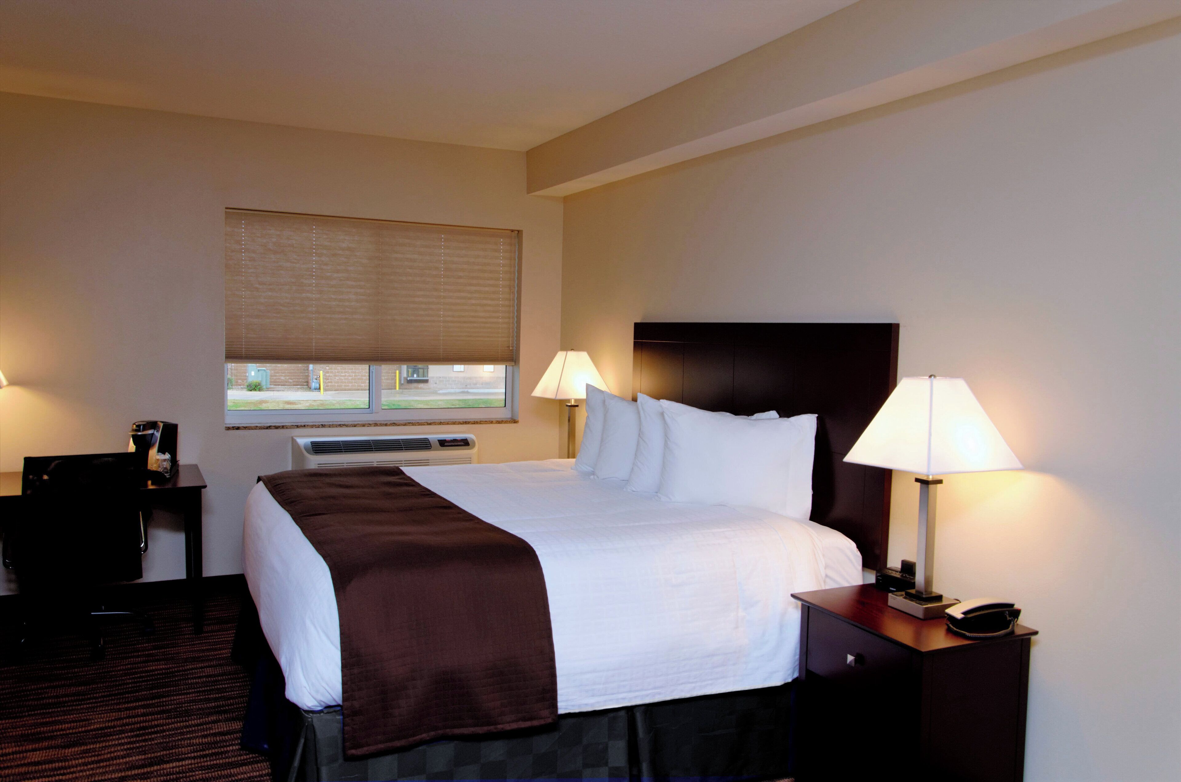 A hotel room with a large bed, two bedside lamps, a desk with a chair, and a window with blinds.