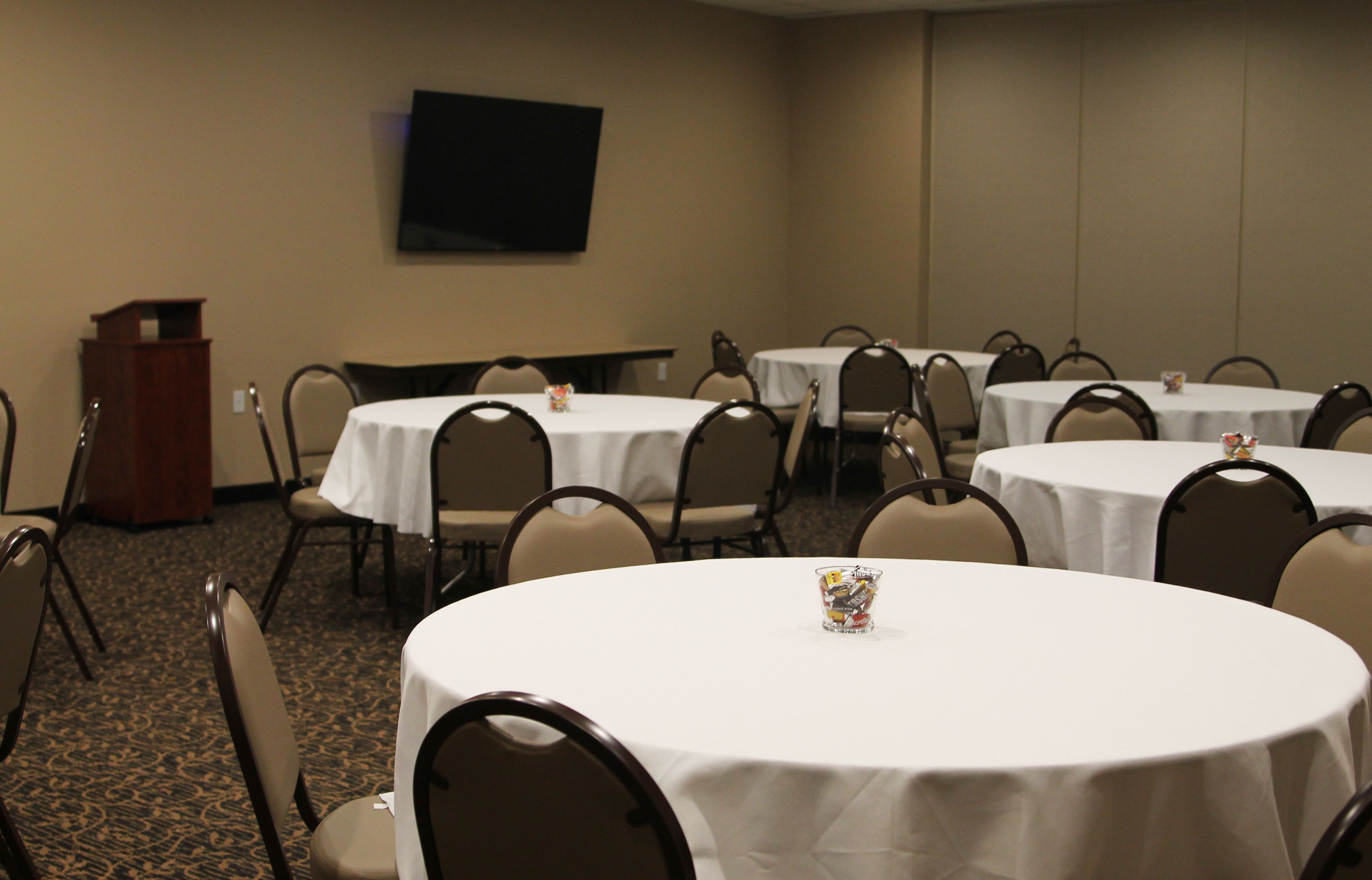 A conference room with multiple round tables, chairs, a podium, and a flat-screen TV on the wall.