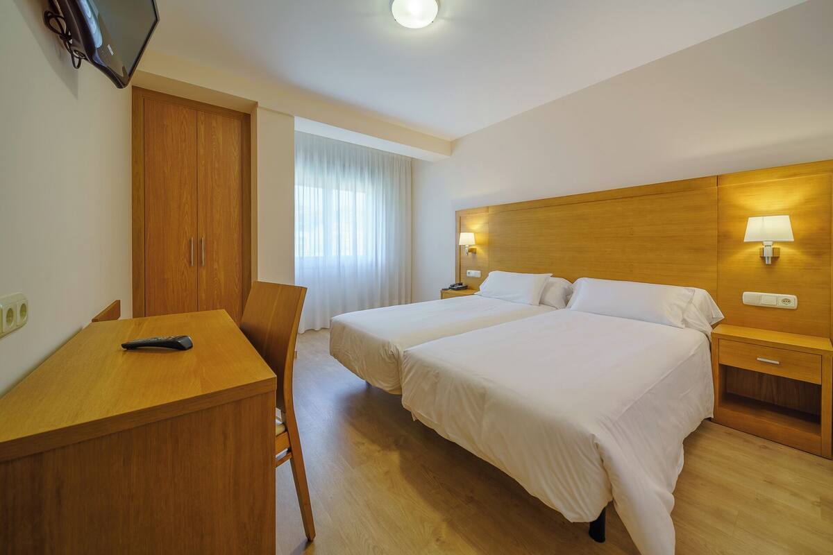 A hotel room with two beds, a desk, a TV, and a window with curtains.