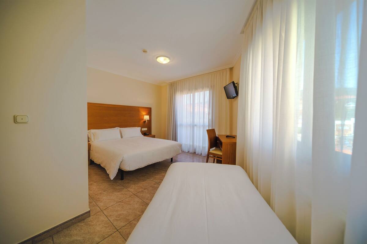 A hotel room with two beds, a desk, a chair, a TV mounted on the wall, and a window with curtains.