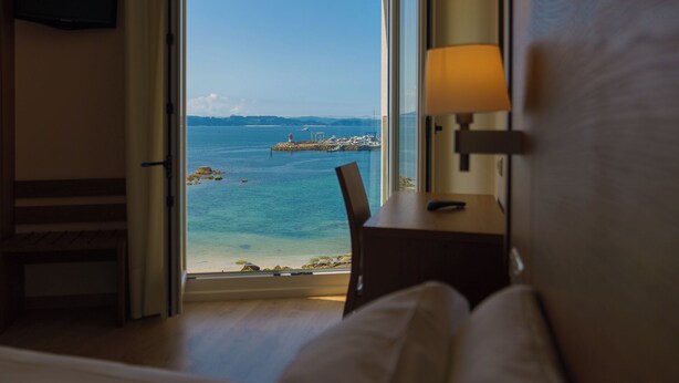 A hotel room with a large window offering a view of the sea, a wooden table, and a lamp. - Hotel El Puente (Sanxenxo)