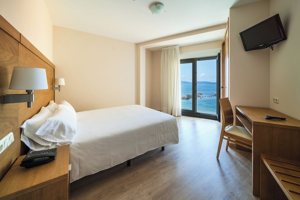 A hotel room with a bed, a desk, a chair, a TV, and a view of the sea.