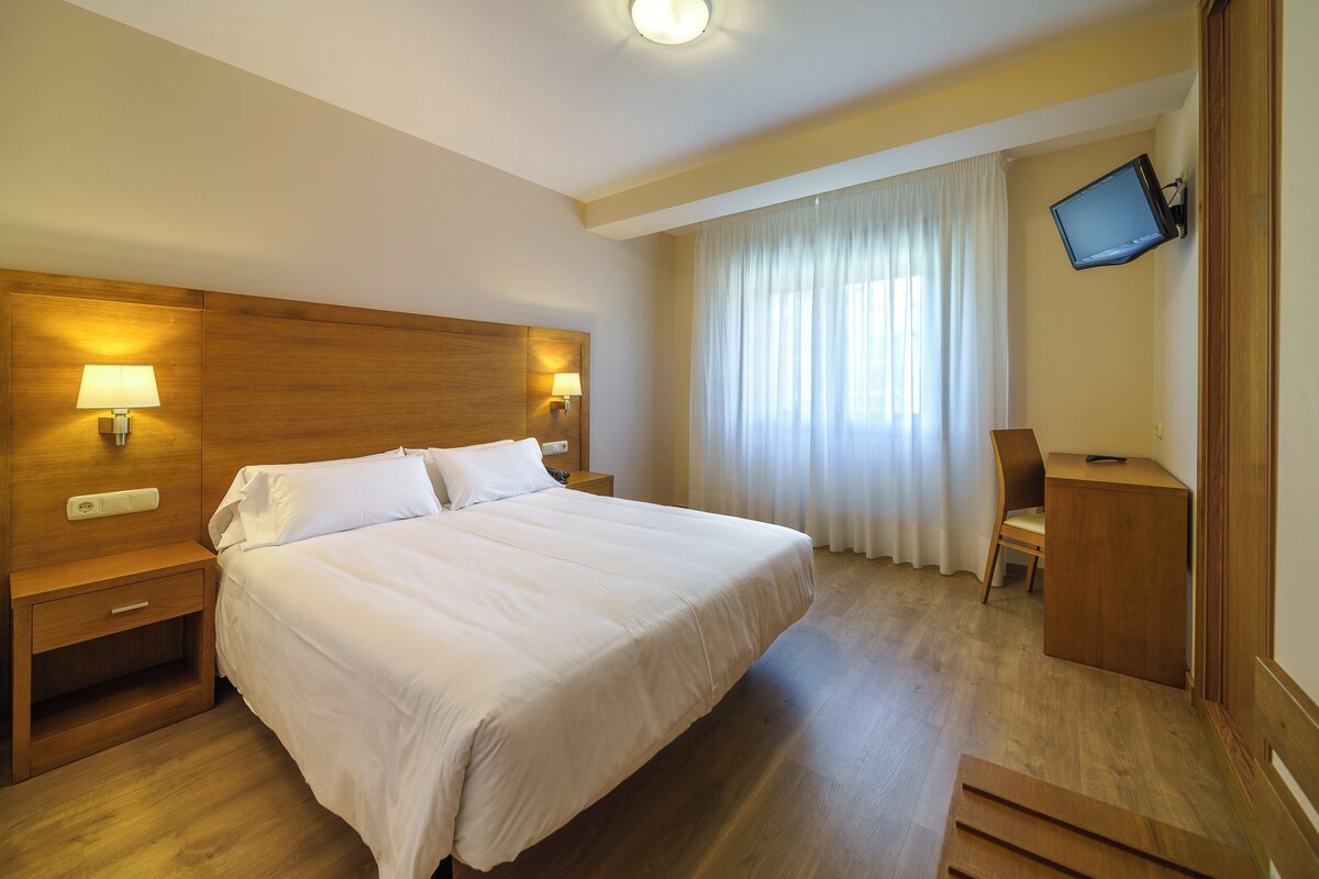 Une chambre d’hôtel dotée d’un grand lit, d’une tête de lit en bois, de lampes de chevet, d’un bureau avec une chaise, d’un téléviseur fixé au mur et d’une fenêtre avec des rideaux.