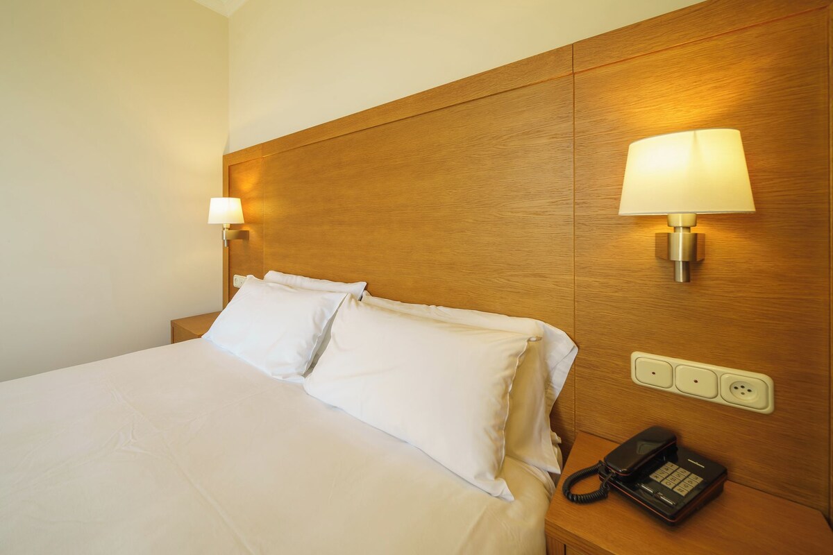 Une chambre d’hôtel avec une tête de lit en bois, deux oreillers blancs, une lampe de chevet, un téléphone et une prise électrique.