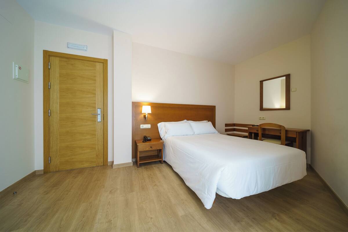 A hotel room with a wooden door, a bedside table with a lamp, a bed, a desk, and a mirror on the wall.