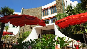 Ein Gebäude mit einer Steinauffachung, ein Restaurant mit rotem Markisenvorzelt und Sonnenschirme draußen.