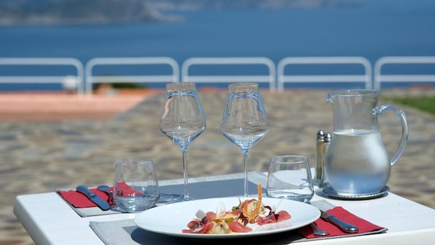 Ein Tisch ist gedeckt mit Essen, Weingläsern und einer Karaffe, mit Blick auf einen Strand, eine Geländer und das Meer im Hintergrund.