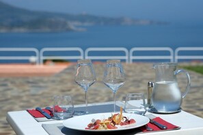 Une table dressée avec une assiette de nourriture, des verres à vin et une carafe, sur une plage avec une rambarde et la mer en arrière-plan.