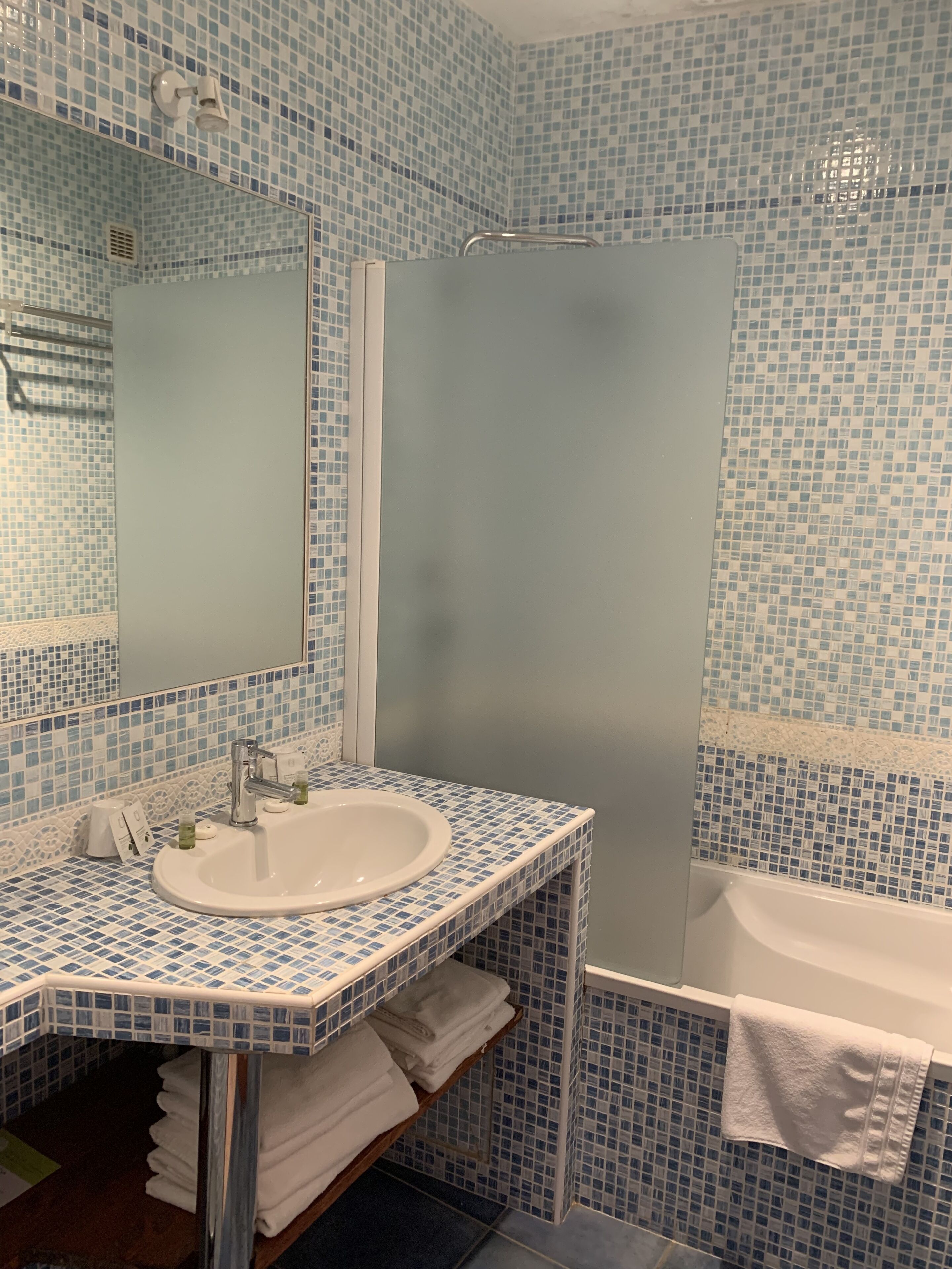 A bathroom with a sink, mirror, and bathtub, all surrounded by blue mosaic tile walls.