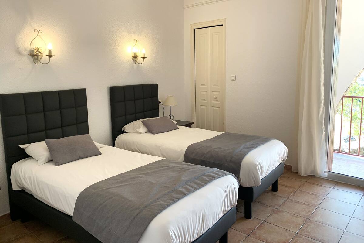 Une chambre d’hôtel avec deux lits, chacun doté d’une tête de lit noire et d’une literie grise. Des lampes murales sont fixées au-dessus des lits, et la porte a un cadre blanc.