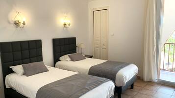 A hotel room with two beds, each with a black headboard and grey bedding. There are wall-mounted lights above the beds and a door with a white frame.