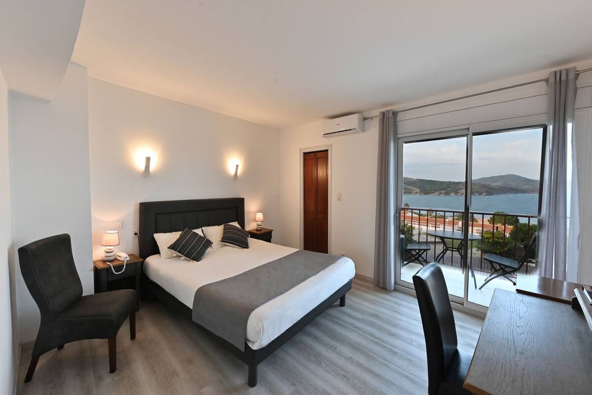 Une chambre d’hôtel avec un grand lit, des tables de chevet, une chaise, un bureau et un balcon offrant une vue sur la mer et les collines.