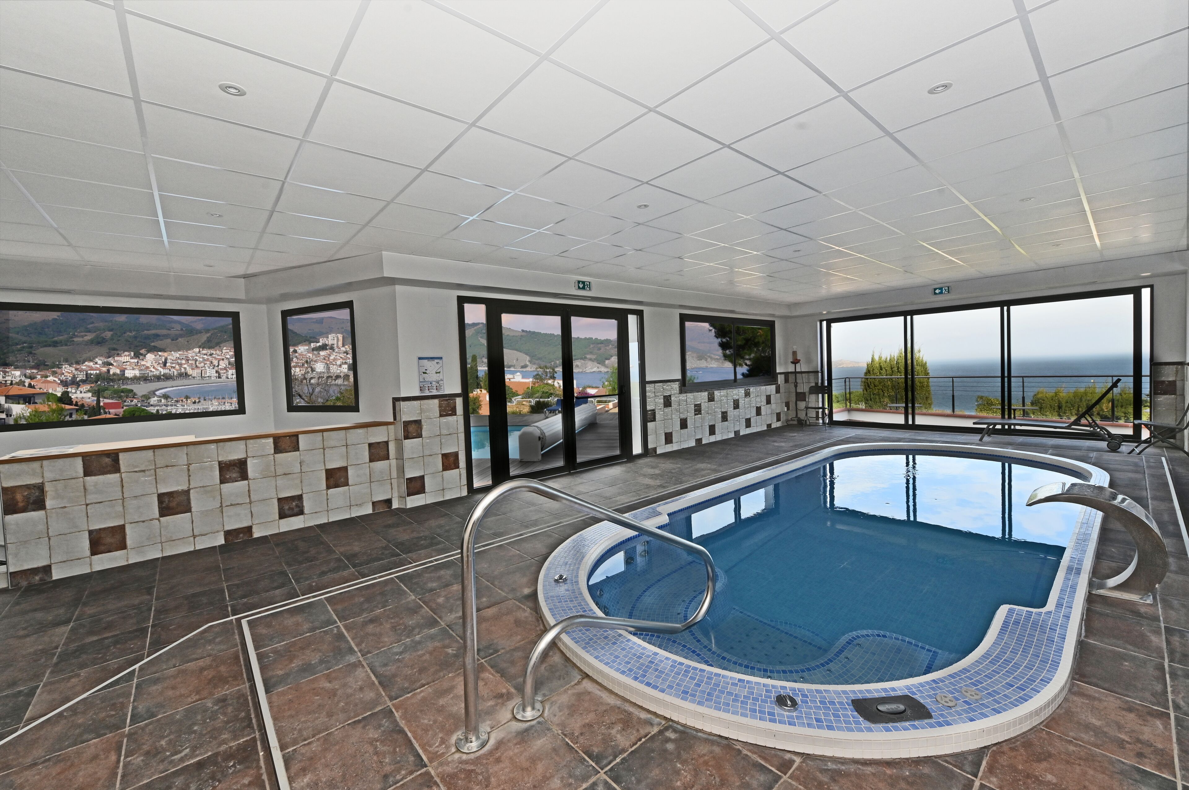A modern indoor swimming pool with a panoramic view of a coastal town.