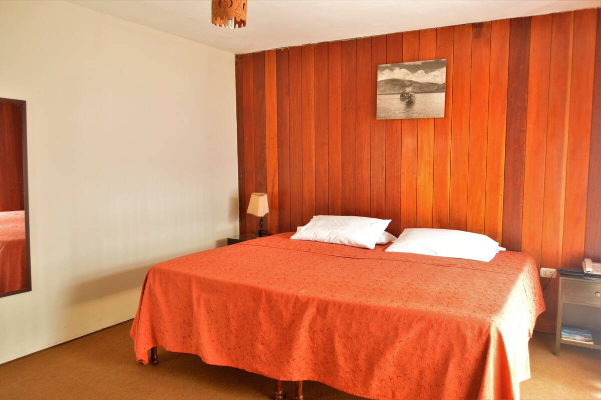 Un dormitorio con un revestimiento de madera, una cama con colcha naranja, una mesita de noche con una lámpara y un cuadro sobre la cama.