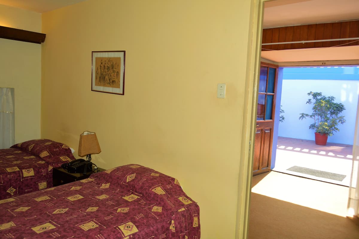 Habitación de hotel con dos camas, una mesita de noche, una lámpara, un cuadro en la pared y vistas al balcón exterior con una planta en maceta.