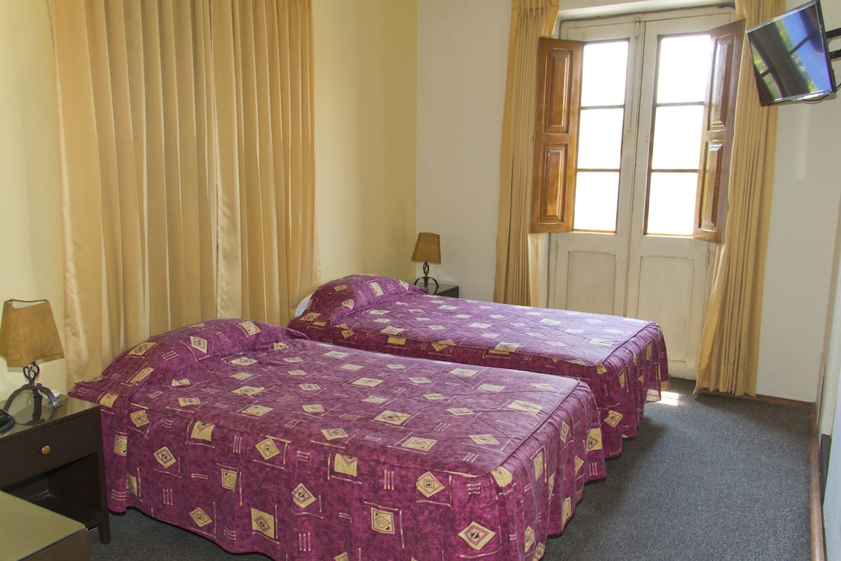 Habitación de hotel con dos camas, un televisor en la pared, un escritorio con lámpara y una ventana con cortinas.
