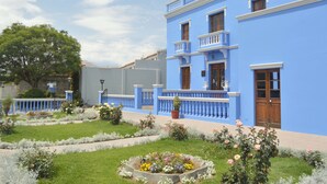 A blue building with a balcony, surrounded by a garden with various flowers and a tree. - Las Mercedes Hostal Residencial (Arequipa)