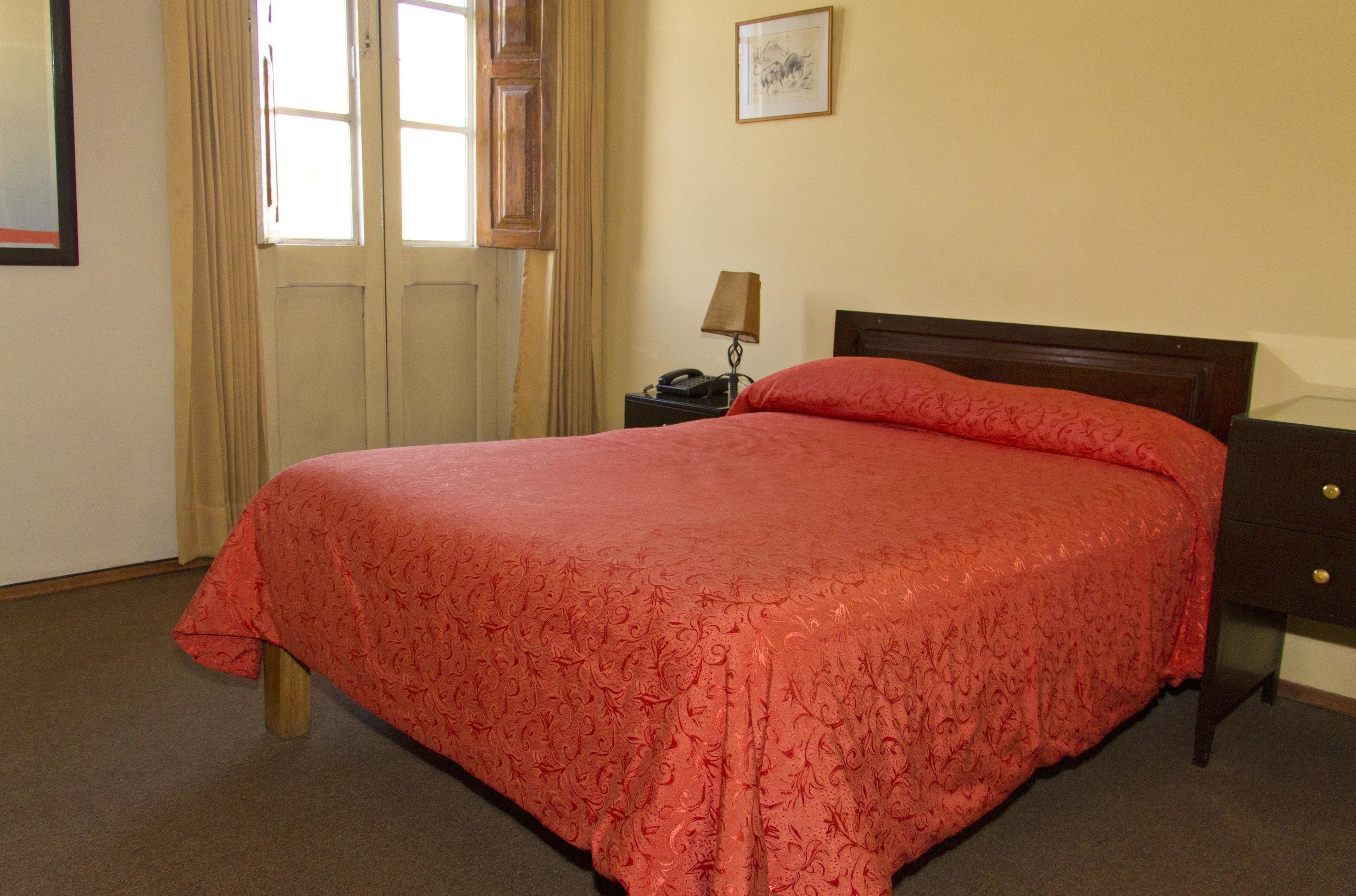A neatly made bed with a red bedspread in a simple room with a wooden headboard, a nightstand with a lamp, and a door with curtains.