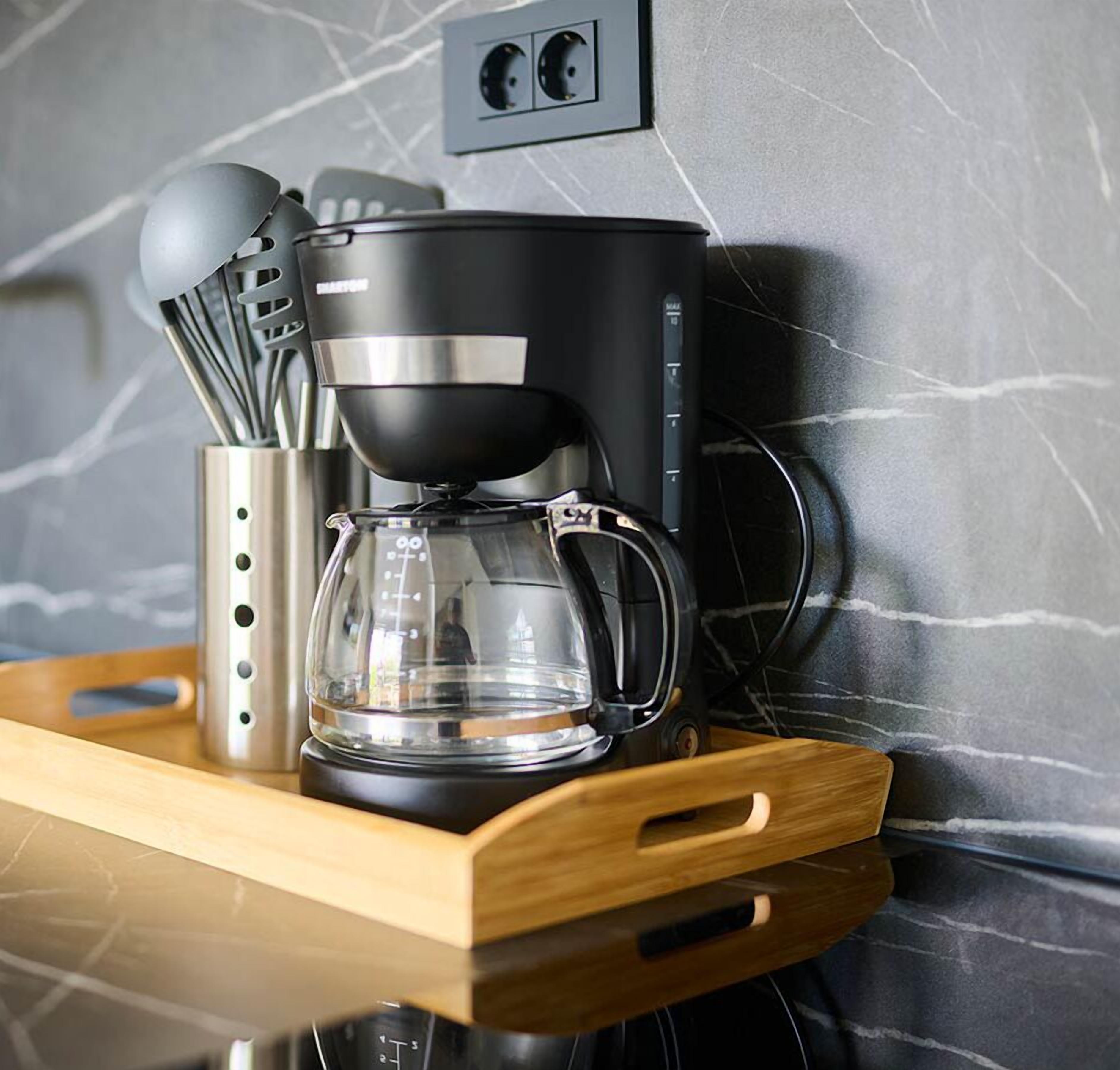 A coffee maker on a wooden tray with a glass carafe, set against a marble wall with a power outlet above.