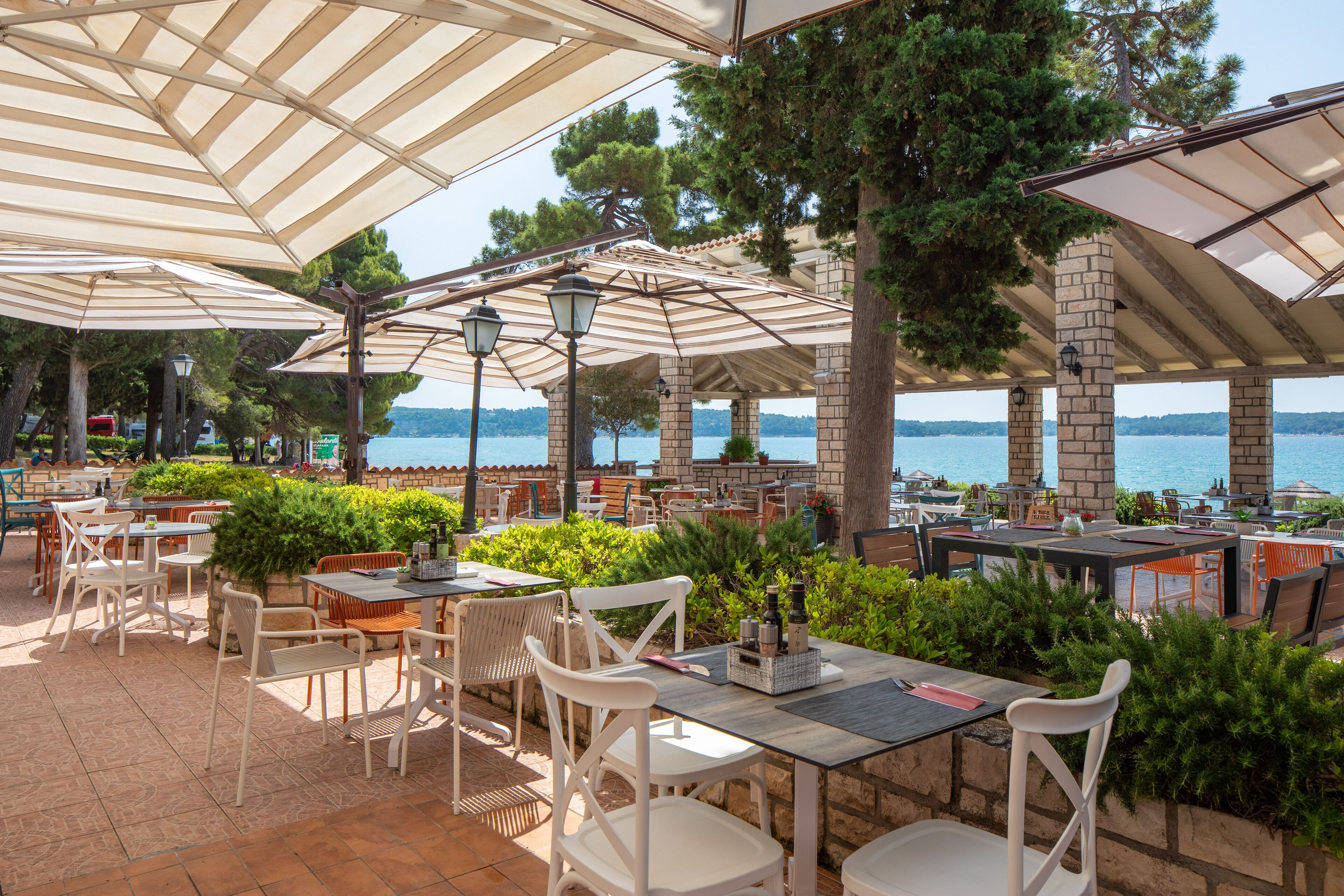 An outdoor patio area with tables and chairs, surrounded by greenery and overlooking a body of water.