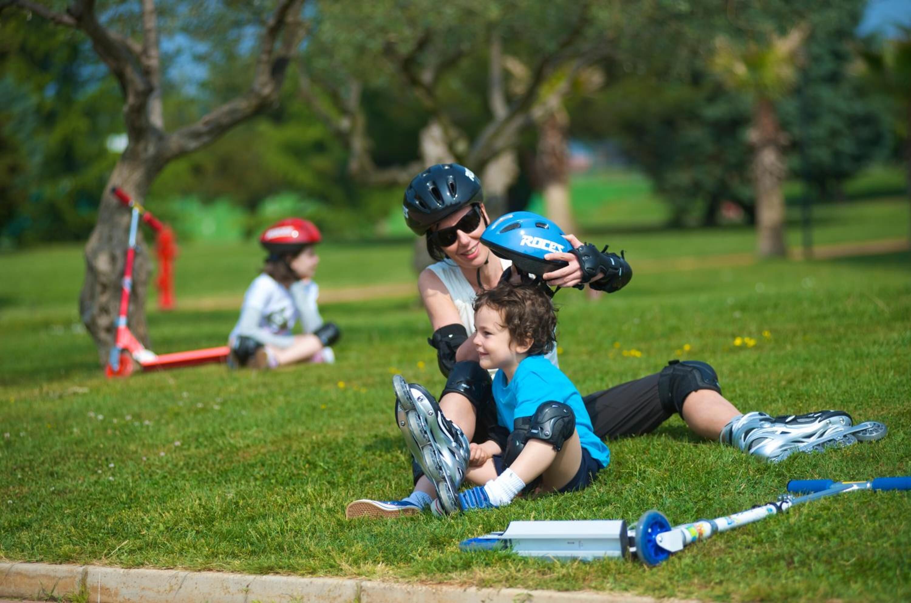 Een grasveld met een persoon in een helm en beschermende kleding, een kind in een blauw shirt, en een rode scooter op de achtergrond.