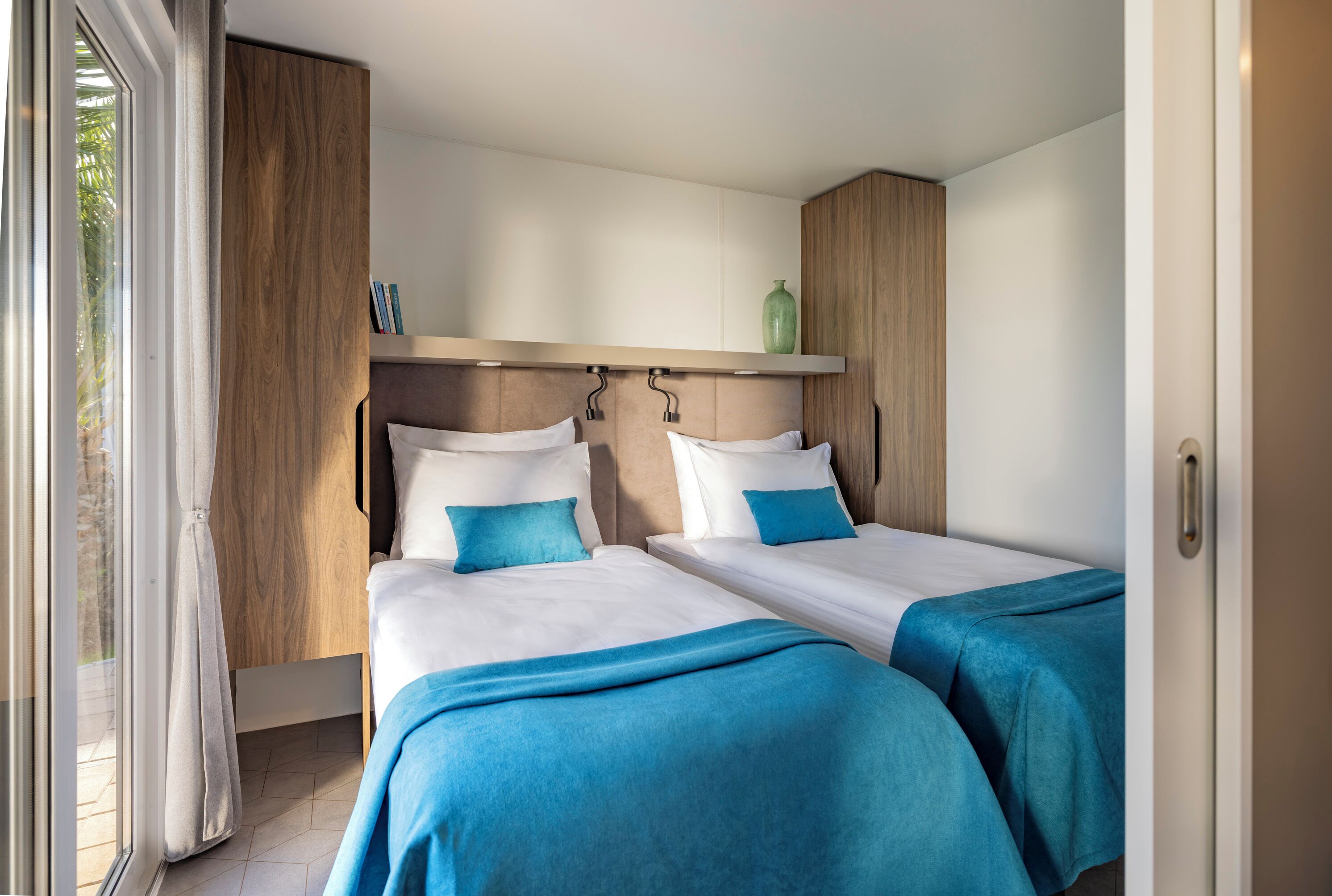 Een kamer met twee bedden, een houten garderobe, een plank met boeken en een groene vaas.