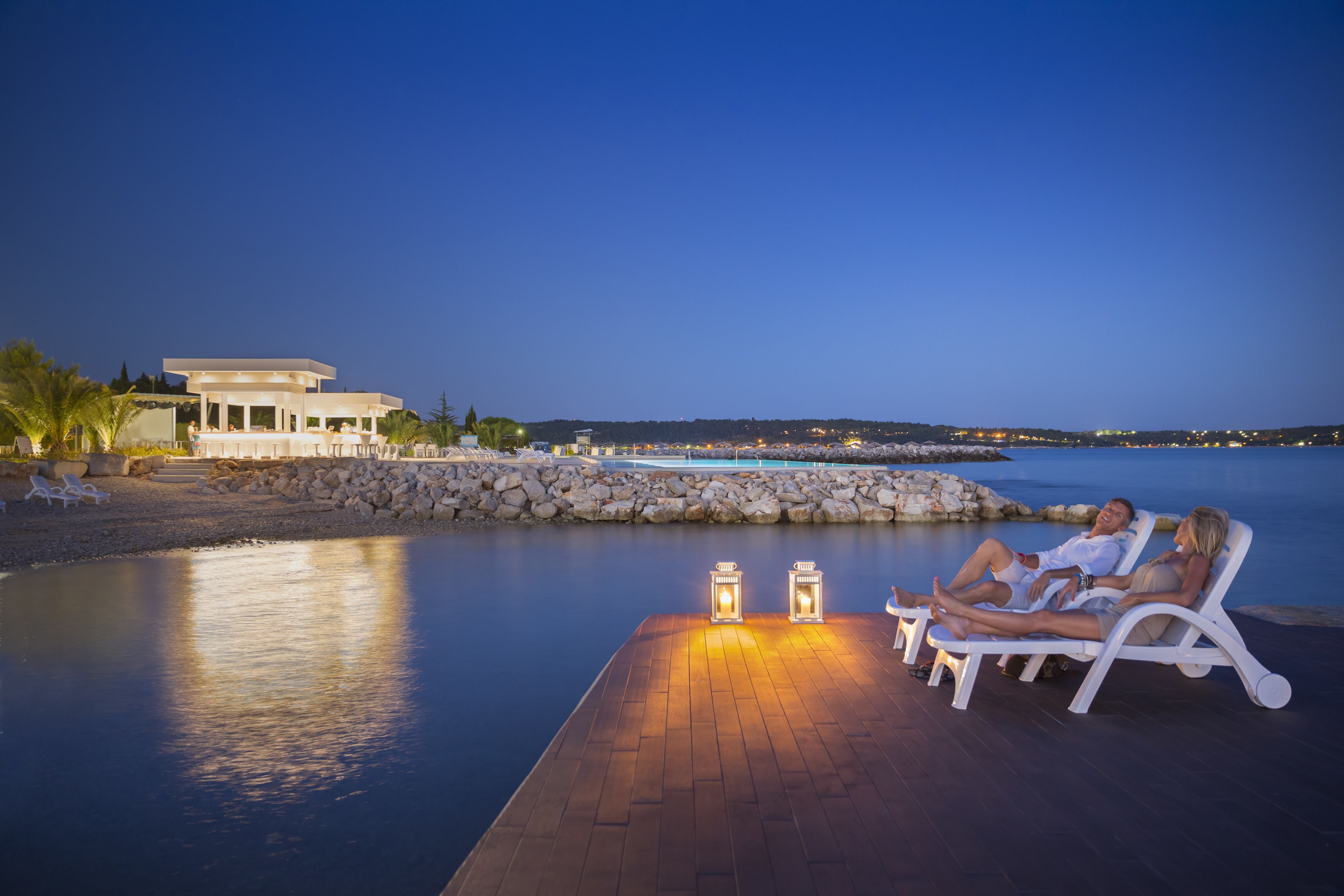 A poolside area with lounge chairs and a wooden deck, overlooking a calm body of water and a distant shoreline.