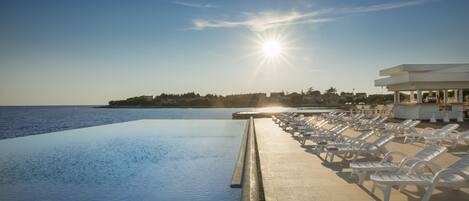 An infinity pool with a view of the sea and a row of white lounge chairs.