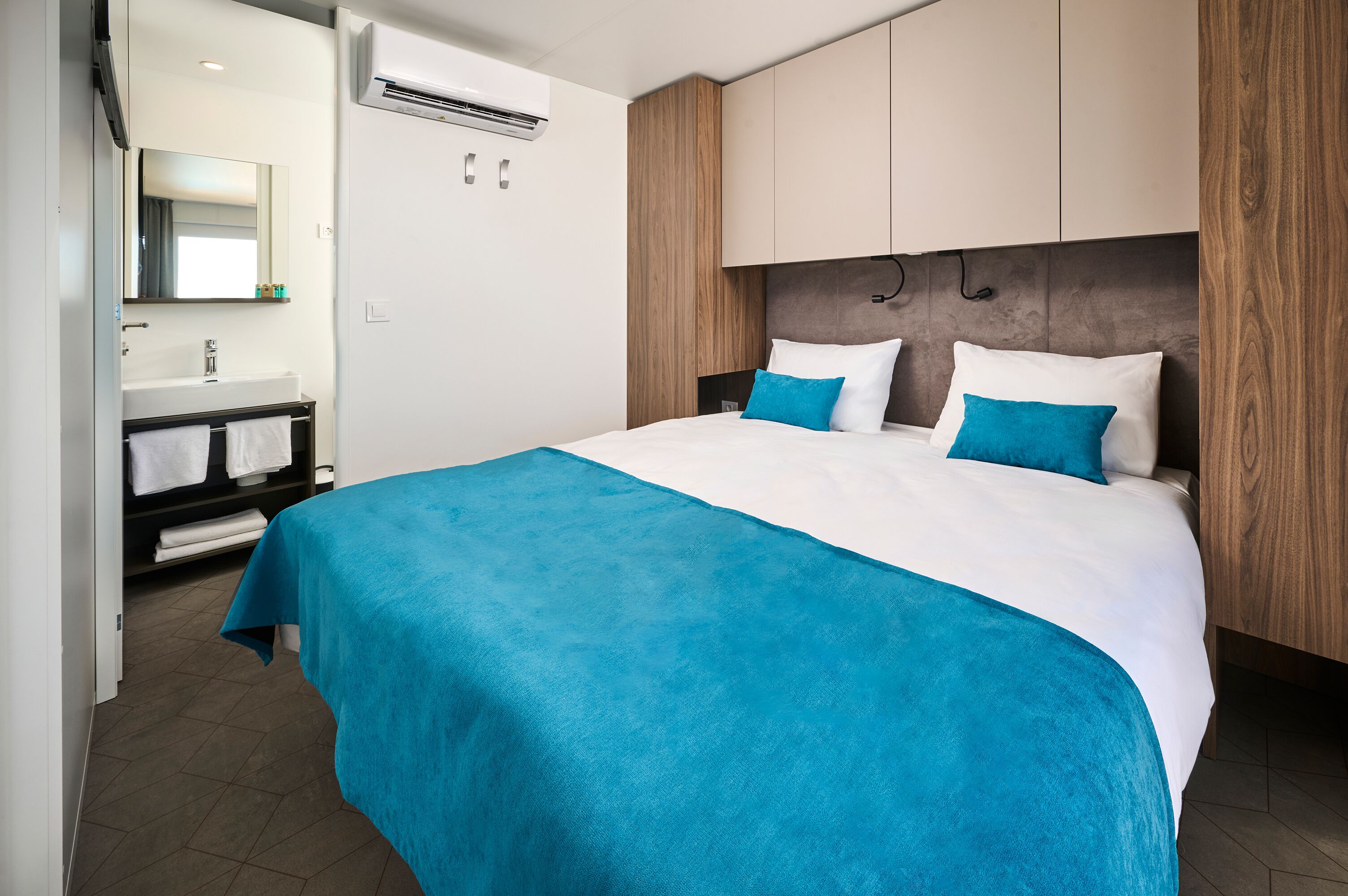 A modern hotel room with a large bed, a built-in wardrobe, a bathroom with a sink and mirror, and a flat-screen TV.