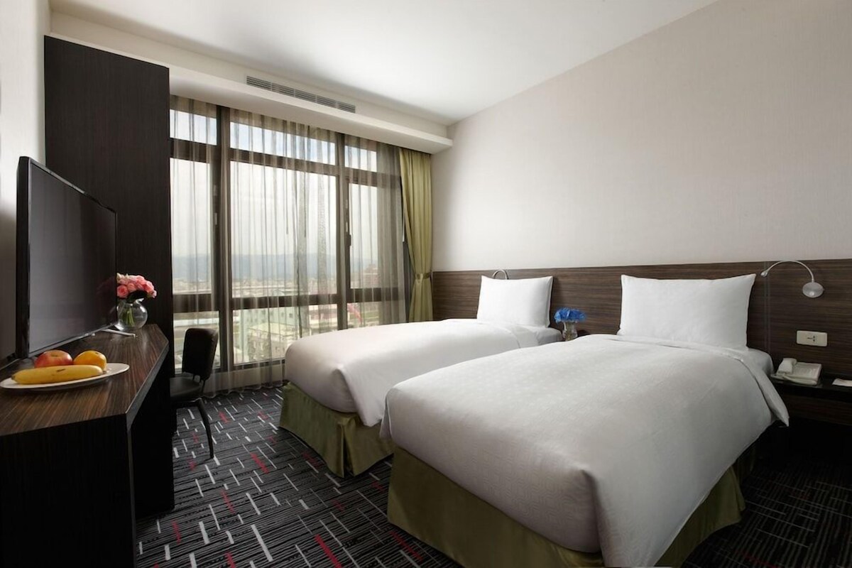 A hotel room with two beds, a desk with fruit, a TV, and a large window with curtains.