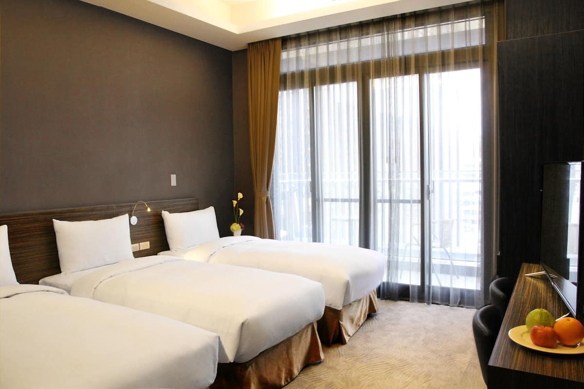 A hotel room with two beds, a desk with a fruit plate, a TV, and a large window with curtains.