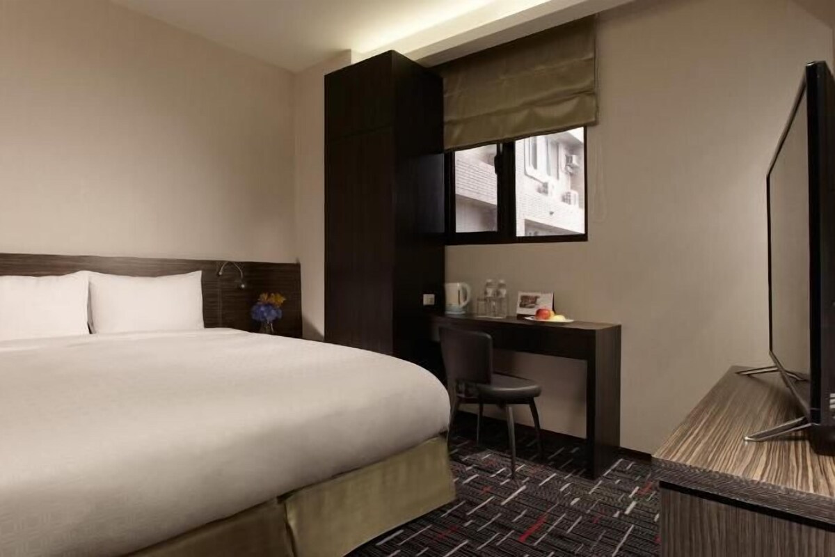 A hotel room with a large bed, a desk, a chair, a TV, and a window with a view.