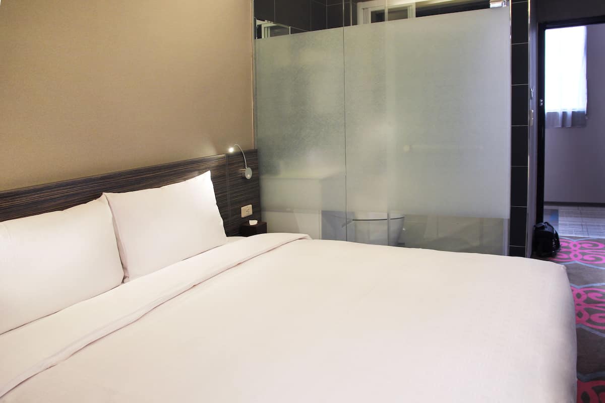 A hotel room with a bed, a nightstand, a glass shower enclosure, and a door leading to another room.